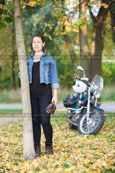 Attractive Caucasian woman biker with a motorcycle is in a park or forest at autumn season, full length portrait. Стоковое фото, фотограф Кекяляйнен Андрей / Фотобанк Лори