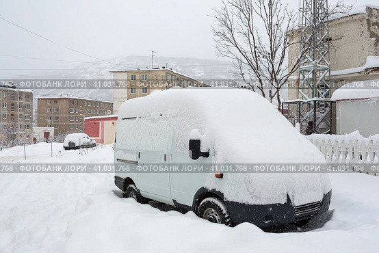 Автомобиль покрыт снегом после снегопада. Сугробы кругом на дороге и во дворе. Снег на крыше и лобовом стекле автомобиля. На заднем плане панельные дома и сопка. Город Магадан, Магаданская область, Россия. Стоковое фото, фотограф Андрей Степанов / Фотобанк Лори