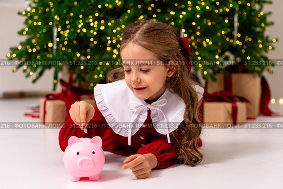 Banking, finances, investment and happy people concept. Cute happy little girl lies a floor and puts money coins in pink piggy bank on Christmas. Pocket money, family budget and savings concept. Стоковое фото, фотограф Ирина Михайличенко / Фотобанк Лори
