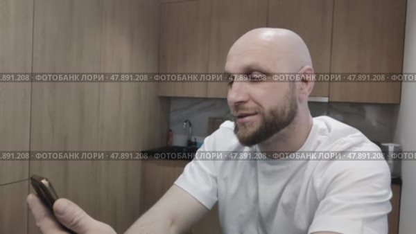 Bearded man talking via video link with his relatives from another city or country. Guy having lively conversation on his smartphone, sitting at home in kitchen. Portrait of young man with phone. Стоковое видео, видеограф Ирина / Фотобанк Лори