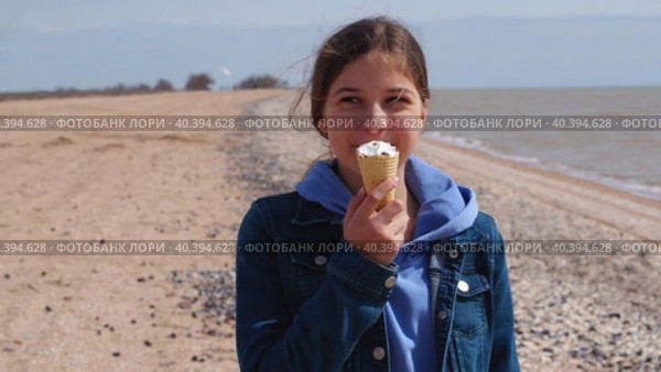 Beautiful girl with an ice cream cone on the beach. A cute teenage girl eats ice cream and smiles while looking at the camera on an empty autumn sea beach. Happy child on the background of the sea. Стоковое видео, видеограф Александр Кротков / Фотобанк Лори