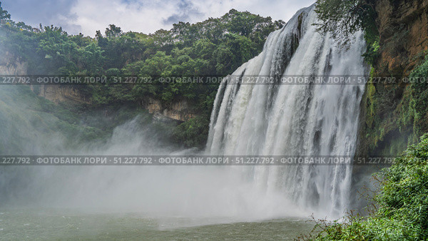 Beautiful tropical waterfall. Streams of water flow down from a sheer cliff into the river. Foam, splashes, fog. Green vegetation on the rock. Clouds in the sky. China. Huangguoshu Waterfall. Guizhou. Стоковое фото, фотограф Вера Ивановна Кубарева / Фотобанк Лори