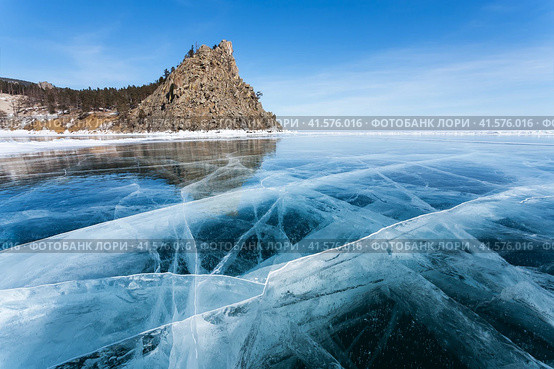 Beautiful winter unusual landscape of frozen Baikal Lake on February sunny day. View from blue ice with cracks of famous sandy beach of Peschanaya bay and  Bell Tower Rock - natural landmark. Стоковое фото, фотограф Виктория Катьянова / Фотобанк Лори