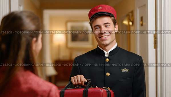 Bellboy carrying luggage smiling at hotel guest. Стоковое AI-изображение, AI-художник Юрий Поздников / Фотобанк Лори