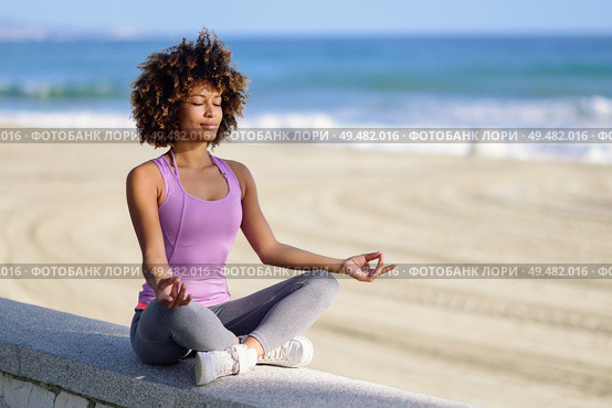Black woman, afro hairstyle, doing yoga in the beach with eyes closed. Young Female wearing sport clothes in lotus figure with defocused background.. Black woman, afro hairstyle, in lotus asana with eyes closed in the beach. Стоковое фото, агентство Ingram Publishing / Фотобанк Лори