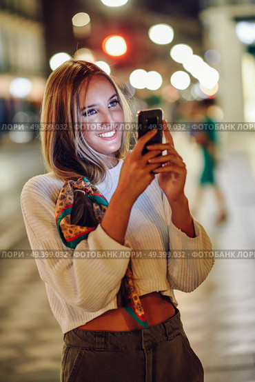 Blonde woman taking photograph with smartphone at night in the street. Defocused city lights at the background. Pretty girl with pigtail hairstyle at night smiling. Стоковое фото, агентство Ingram Publishing / Фотобанк Лори