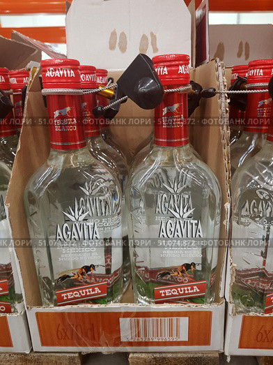 Bottles of Agavita Blanco tequila neatly arranged in boxes on a store shelf. Clear glass bottles with a bright red cap and a protective anti-theft device. The label depicts agave, a symbol of the drink's main ingredient, as well as inscriptions in Spanish indicating its origin in Mexico. At the bottom of the label is the word Tequila on a red background. The crystal clear contents are visible through the clear glass. In the background are repeating rows of bottles, creating a rhythmic composition. (2019 год). Редакционное фото, фотограф Анна Кривицкая / Фотобанк Лори