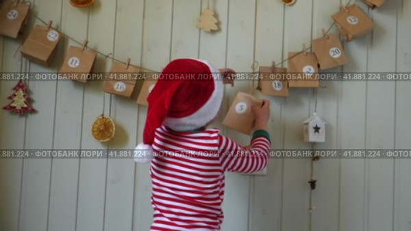 Boy in pajamas, Santa cap eating sweet candy on paper advent calendar with presents background. Little happy child with gift of handmade Christmas calendar on wall. Celebrating at home Xmas tradition. Стоковое видео, видеограф Марина Демидюк / Фотобанк Лори