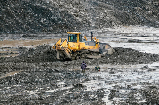 Бульдозер Komatsu D375A в горной местности, снимает верхний слой грунта. Рядом опробщик ( старатель-разведчик ), который контролирует с помощью промывочного лотка концентрацию золотых частиц в горном грунте (2009 год). Редакционное фото, фотограф Павел Просянов / Фотобанк Лори