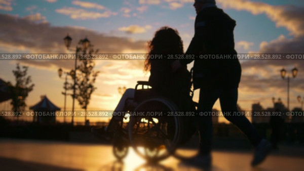 Caring man with a disabled woman in wheelchair walking through the quay at sunset. Стоковое видео, видеограф Константин Шишкин / Фотобанк Лори