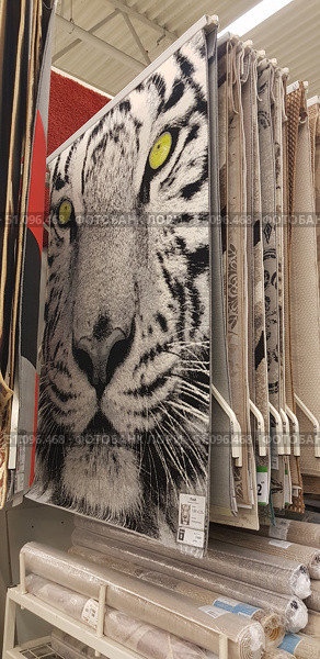 Carpet and flooring store. The center of the frame features a carpet with a bright and striking design of a white tiger's head with yellow eyes. The black and white image of the predator creates a contrasting and expressive impression, making the product a decorative accent. The carpets are hung vertically for display, with other models with traditional and modern patterns in calm tones visible nearby. Rolled carpets and rugs are located on a shelf below. The scene conveys the atmosphere of the home interior department in a home goods hypermarket. (2019 год). Редакционное фото, фотограф Анна Кривицкая / Фотобанк Лори