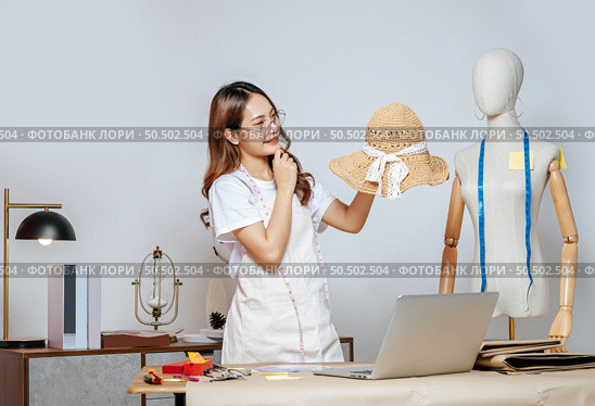 Cheerful Asian young female designer wearing eyeglasses doing fashion design on beautiful sombrero in studio, full of tailoring tools  with mannequin and equipment on the desk. Стоковое фото, агентство Ingram Publishing / Фотобанк Лори