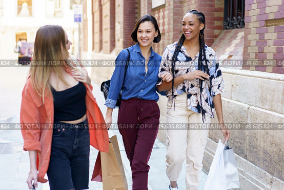 Cheerful multiracial female friends with shopping bags looking at each other while strolling together on walkway near building in city. Smiling diverse women with shopping bags walking on street. Стоковое фото, агентство Ingram Publishing / Фотобанк Лори
