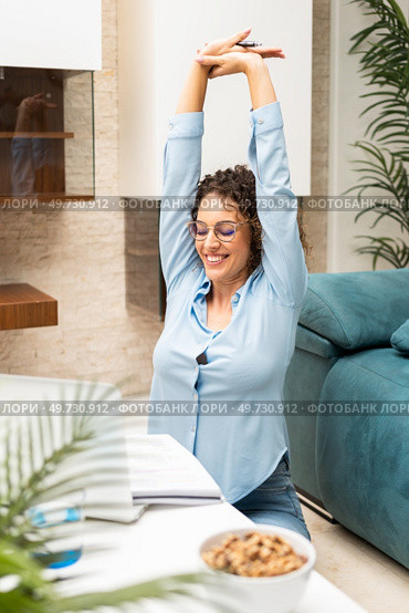 Cheerful young female freelancer in smart casual stretching arms while working on laptop at table in living room at home. Smiling tired woman stretching near netbook at home. Стоковое фото, агентство Ingram Publishing / Фотобанк Лори