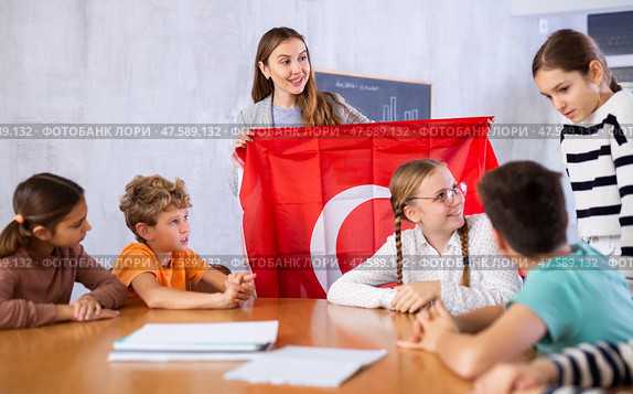 Cheerful young woman teacher demonstrating Turkish flag for schoolkids preteens during lesson of geography in auditory. Стоковое фото, фотограф Яков Филимонов / Фотобанк Лори