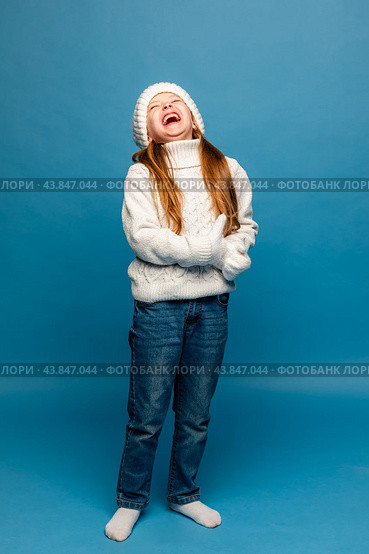 Child girl in a white sweatshirt and knitted hat with pompom on a blue background. The concept of children's emotions, joy and delight. Advertising clothing and sales. Ski resort vacation, copy space. Стоковое фото, фотограф Ирина Михайличенко / Фотобанк Лори