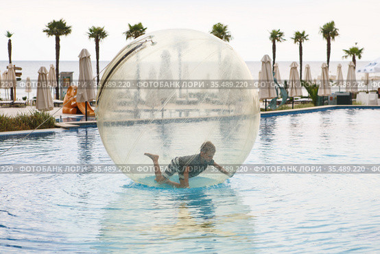 Child have fun inside big plastic balloon on the water of swimming pool on the summer resort. Little boy inside big inflatable transparent ball. Стоковое фото, фотограф Nataliia Zhekova / Фотобанк Лори