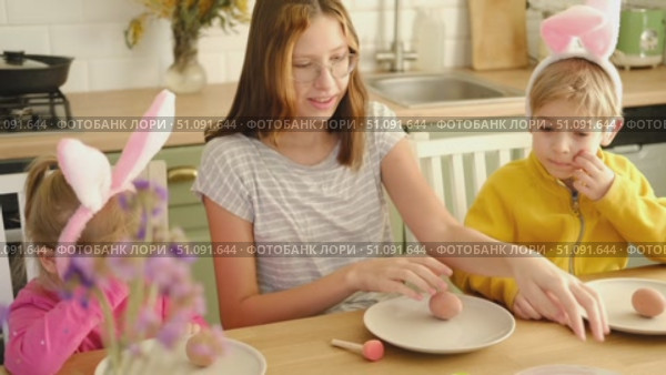 Children siblings painting Easter egg sitting together at table. Easter Family traditions. Teen girl teaching happy little brother and sister to dye, decorate eggs with paints. Preparation to holidays. Стоковое видео, видеограф Марина Демидюк / Фотобанк Лори