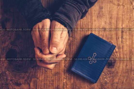 Christian man praying with hands folded and fingers crossed with Holy Bible by his side on wooden desk in church, top view. Стоковое фото, агентство Ingram Publishing / Фотобанк Лори