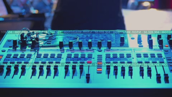 Close-up of a mixing console illuminated by blue light. Various switches, knobs and buttons are visible, showing a professional setup. There is a live performance in the background. Стоковое видео, видеограф Воловодов Владимир / Фотобанк Лори