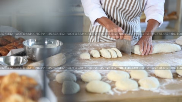 Close-up of skilled baker hands cutting soft dough with scraper on floured wooden worktable, filled with baking tools and ingredients, in bakery. Стоковое видео, видеограф Яков Филимонов / Фотобанк Лори