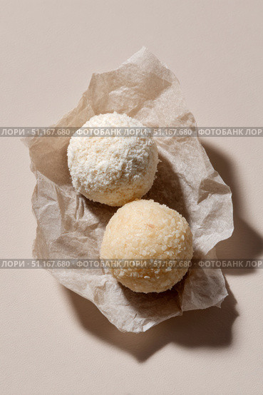 Coconut candy spheres with contrasting texturesone rolled in fine shredded coconut, another in larger flakespositioned on crinkled parchment over neutral background. Стоковое AI-изображение, AI-художник Юрий Поздников / Фотобанк Лори