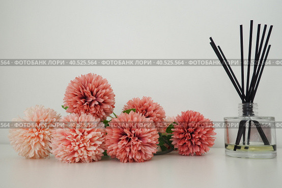 Composition with incense sticks, essential oil in a glass vase and pink terry artificial flowers in the interior of a white room. White background. Lots of flowers and scent sticks. Стоковое фото, фотограф Юлия Петровская / Фотобанк Лори