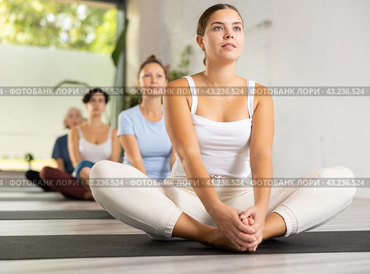 Concentrated young woman performing Butterfly Pose Baddha Konasana during yoga class with female group. Стоковое фото, фотограф Яков Филимонов / Фотобанк Лори