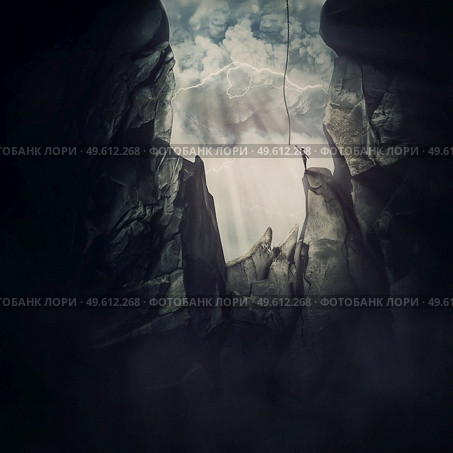 Conceptual scene with a man escaping from the steep chasm, stretching to reach the helping rope and get out of the darkness. Big cave inside the rocky mountain and person trying to break free. Стоковое фото, агентство Ingram Publishing / Фотобанк Лори