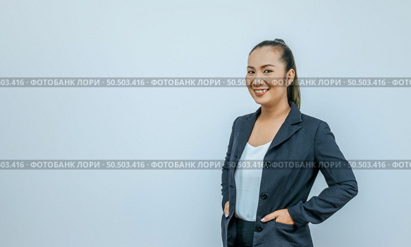 Confident Asian woman smiling, looking at camera posing over  background with blank space. Studio portrait of successful friendly female in blazer, keeping hands in pockets. Стоковое фото, агентство Ingram Publishing / Фотобанк Лори