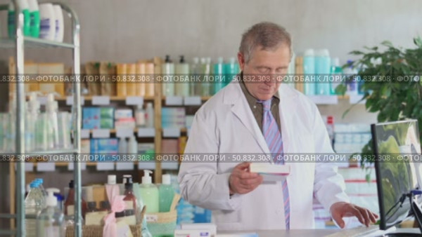 Confident professional senior pharmacist in white coat working behind drugstore counter, inputting purchase orders into computer system, managing incoming supplies and ensuring stock availability. Стоковое видео, видеограф Яков Филимонов / Фотобанк Лори