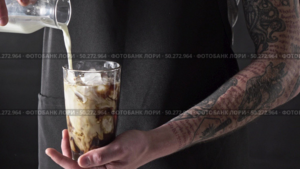 Cooking summer drink. A man with a tattoo pours milk into a glass with coffee and ice around a dark background. 4K UHD video, 240fps, 2160p.. A male barista with a tattoo pours fresh milk into a glass with ice and coffee around a dark background. Cooking Frappe. 4K UHD video, 2160p. Стоковое фото, агентство Ingram Publishing / Фотобанк Лори