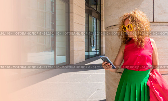 Coquettish s young female model in fashionable elegant bright multicolored green and red clothes looks at her phone against the background of the columns of an office building in the city. Стоковое фото, фотограф Anastasia Kayander / Фотобанк Лори