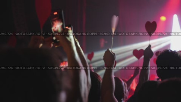 Crowd of concertgoers holding red heart-shaped signs under vibrant stage lights. Стоковое видео, видеограф Данил Руденко / Фотобанк Лори