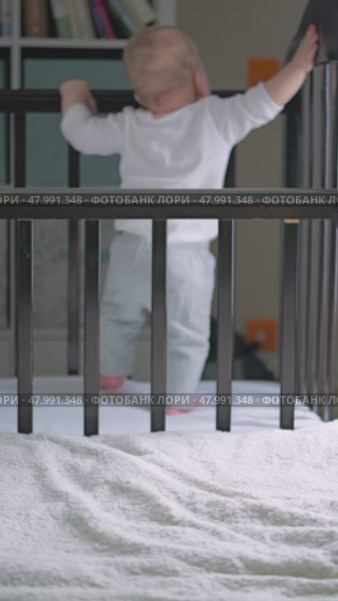 Curious baby stands in the crib, holding on to the edge, dressed in gray pants and a white shirt. The toddler takes his first steps with support. Focus on the crib railing. Стоковое видео, видеограф Воловодов Владимир / Фотобанк Лори