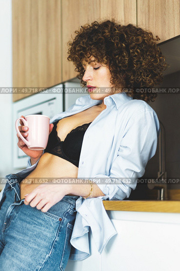 Curly haired woman in black bra unbuttoned shirt and unzipped jeans leaning on counter while enjoying hot beverage. Woman with unbuttoned shirt drinking coffee in kitchen. Стоковое фото, агентство Ingram Publishing / Фотобанк Лори