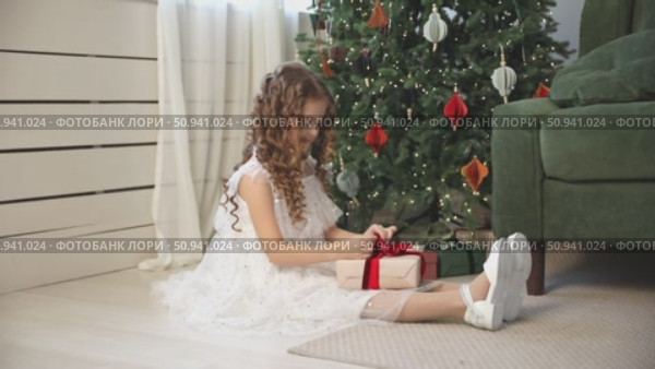 Cute happy little girl with curly hair, in an elegant white fluffy dress sits on floor under decorated Christmas tree at home, joyfully opening a gift box. Childhood and happy family holidays concept. Стоковое видео, видеограф Ирина Михайличенко / Фотобанк Лори