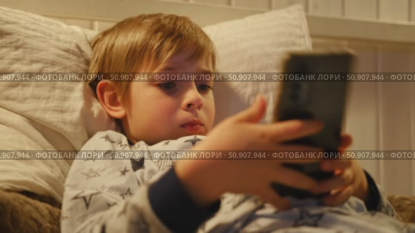 Cute kid boy in pajamas lying in bed watching smartphone, smiling. Children's screen time. Child plays games using phone at the evening or night. Social media addiction. Childhood, modern technology. Стоковое видео, видеограф Марина Демидюк / Фотобанк Лори