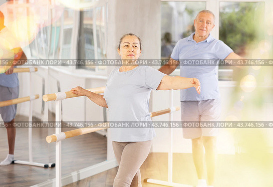 Dancing positive elderly women and men engaged in group class perform exercise near ballet barre, standing in 2nd position of ballet stance in dance studio. Стоковое фото, фотограф Яков Филимонов / Фотобанк Лори