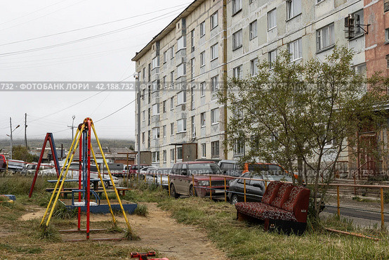 Детские качели и диван на детской площадке в городском дворе возле старого панельного дома. Необустроенное место отдыха возле жилого дома. Проблемы благоустройства старых городских кварталов. Магадан, Магаданская область, Россия. (2021 год). Стоковое фото, фотограф Андрей Степанов / Фотобанк Лори