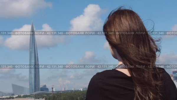 Девушка с каштановыми волосами стоит спиной и смотрит вдаль на  Финский залив и башню Лахта-центра в Санкт-Петербурге. Редакционное видео, видеограф Глущенко Елена / Фотобанк Лори