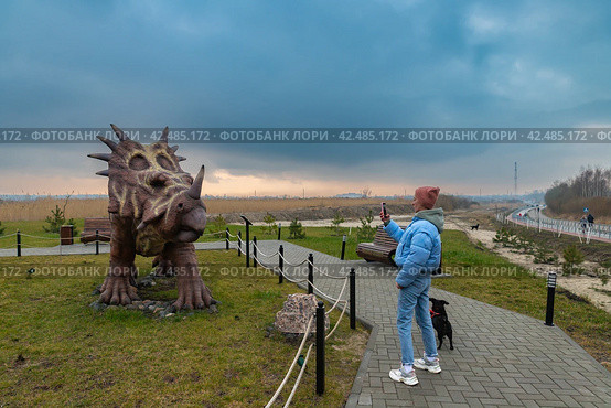 Девушка с собакой фотографирует фигуру стиракозавра в Динопарке Янтарного (2024 год). Редакционное фото, фотограф Константин Чижов / Фотобанк Лори