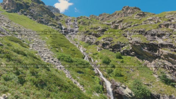 Длинный водопад каскадом стекает по скалам в высоких горах. Длинный водопад талой воды. Панорама сверху вниз. Скалы вокруг покрыты зеленой травой. Горный туризм. Стоковое видео, видеограф Воловодов Владимир / Фотобанк Лори