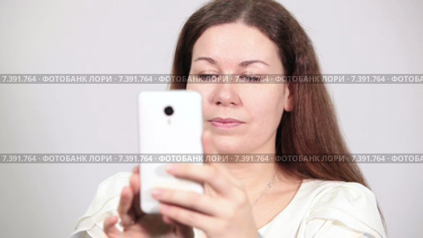 Довольная женщина фотографирует на камеру смартфона, смотрит в экран и нажимает кнопку, серый фон. Стоковое видео, видеограф Кекяляйнен Андрей / Фотобанк Лори