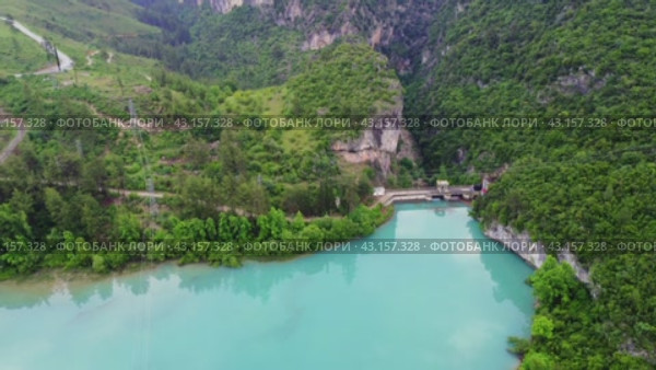 Drone view of mountain turquoise lake and dam. Dam spans lake, highlighting water management. Turquoise lake reflects surrounding landscape. Dam interaction with lake is evident (2024 год). Редакционное видео, видеограф ILIA GOROKHOV / Фотобанк Лори