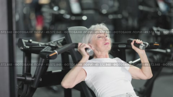 During training session in mini group of athletes, senior woman exercises on shoulder press machine. Стоковое видео, видеограф Яков Филимонов / Фотобанк Лори