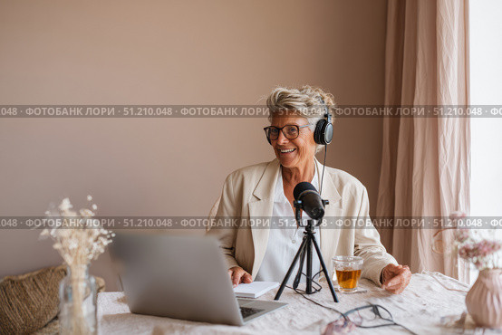 Elderly eastern woman hosting podcast in cozy home with laptop and microphone. Стоковое AI-изображение, AI-художник Евгения Пархоменко / Фотобанк Лори