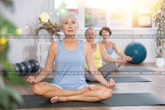 Elderly woman in company of hatha yoga lovers performs half-lotus pose, Ardha Padmasana. Стоковое фото, фотограф Яков Филимонов / Фотобанк Лори