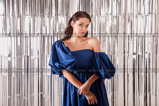 Elegant vogue woman model in blue dress at bright tinsel background celebrating festive holiday, looking at camera. Beautiful positive charming lady posing on glittered backdrop. Copy text space. Стоковое фото, фотограф Aleksandr Golubev / Фотобанк Лори