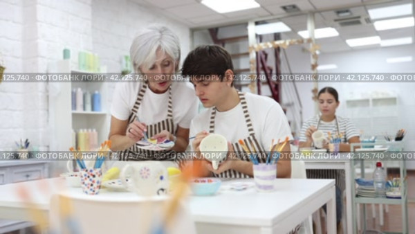 Experienced mature female ceramicist teaching young enthusiastic guy art of ceramic glazing and painting in modern pottery studio. Стоковое видео, видеограф Яков Филимонов / Фотобанк Лори